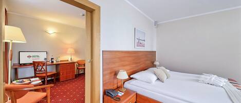 Habitación de hotel con cama, mesita de noche, lámpara, escritorio con computadora, silla y una pequeña mesa con un plato de comida.