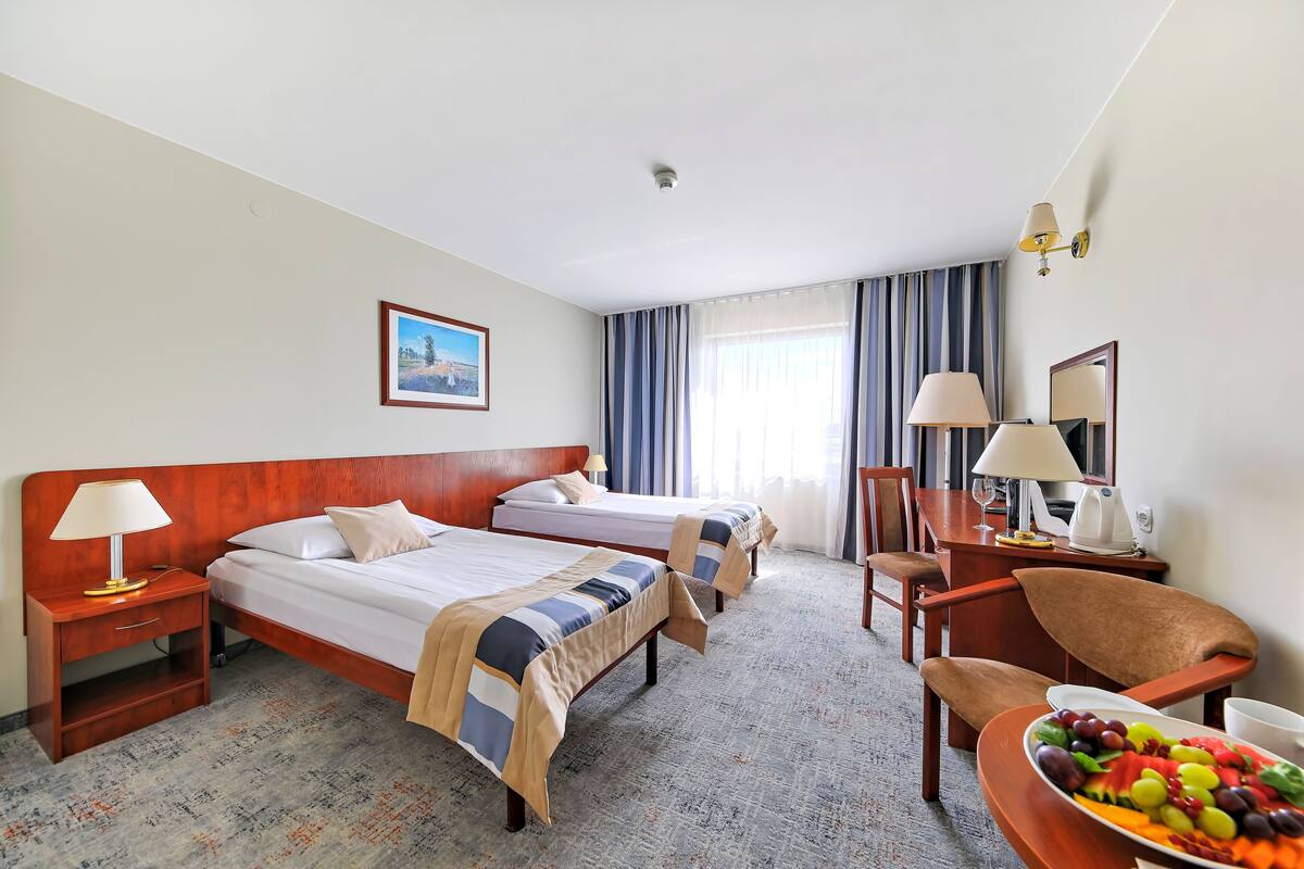 A hotel room with two beds, a desk with a chair, a dining table with a fruit platter, and a painting on the wall.