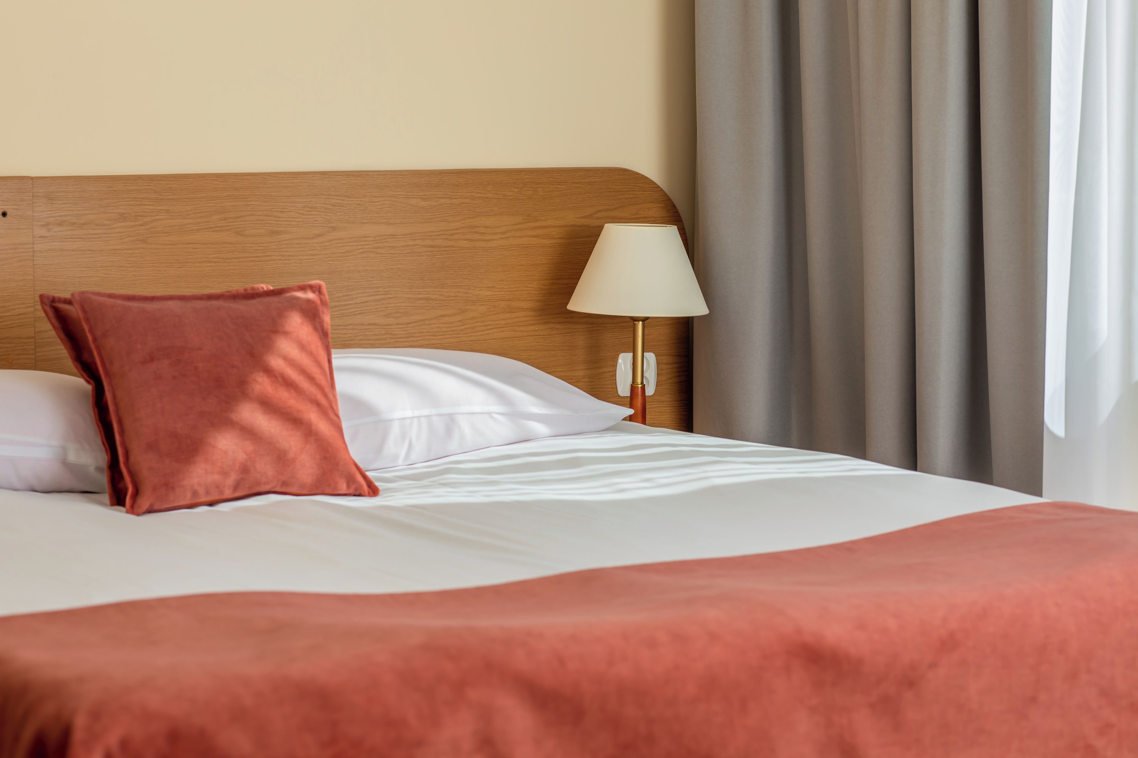 Een hotelkamer met een houten hoofdbord, een enkel bed met witte beddengoed, een rode plaid, een wit kussen en een bedlampje.