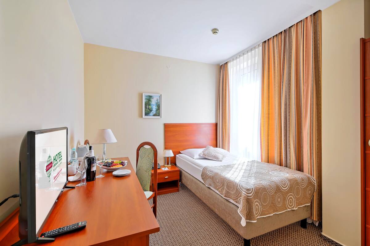 A hotel room with a bed, a desk with a computer monitor, a chair, a nightstand with a lamp, and a window with curtains.