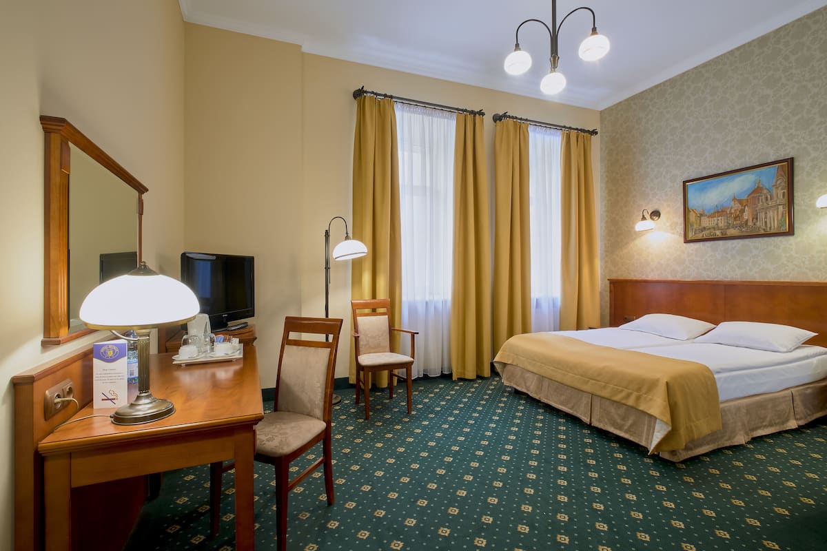 A hotel room with a bed, two chairs, a desk with a lamp, a television, and a painting on the wall.