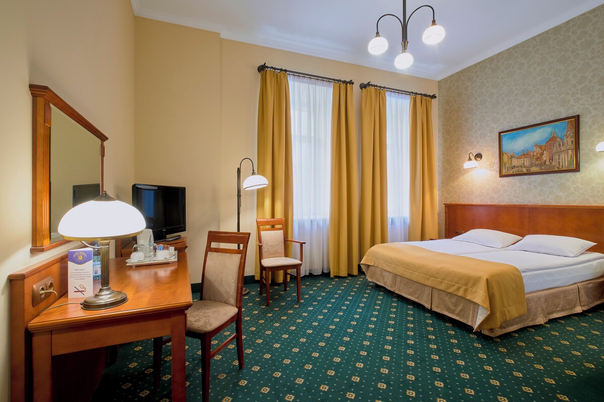 A hotel room with a bed, desk, chairs, a television, and a painting on the wall.