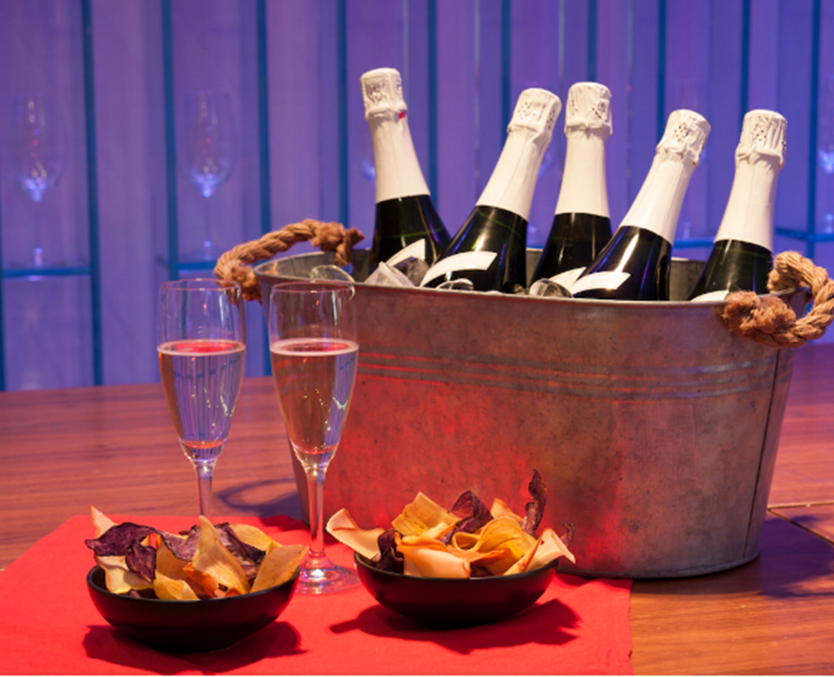 A bucket of champagne bottles, two glasses of champagne, and bowls of snacks on a table.