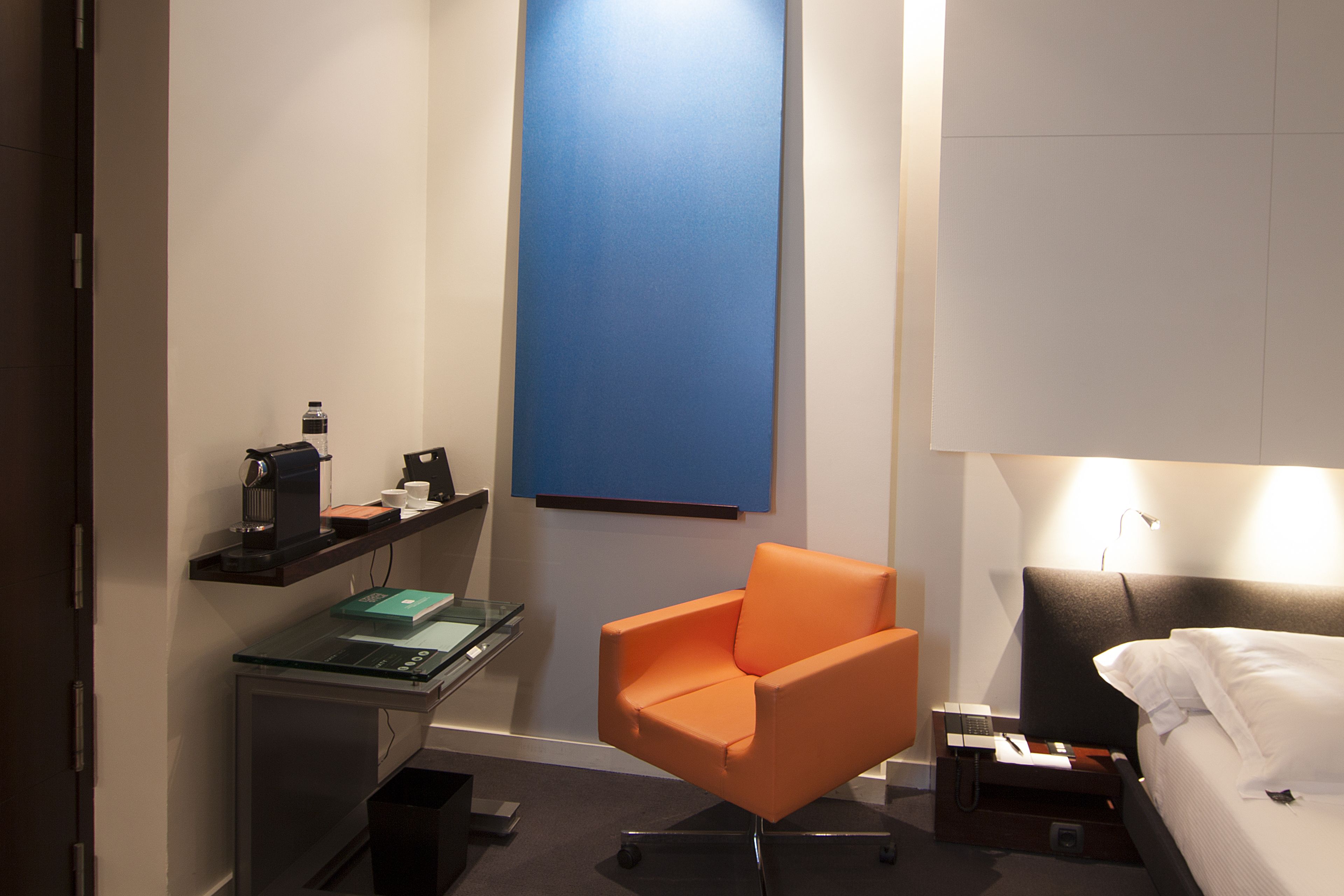 Une chambre d’hôtel avec un lit, un bureau avec un ordinateur, une chaise orange et un mur d’accent bleu.