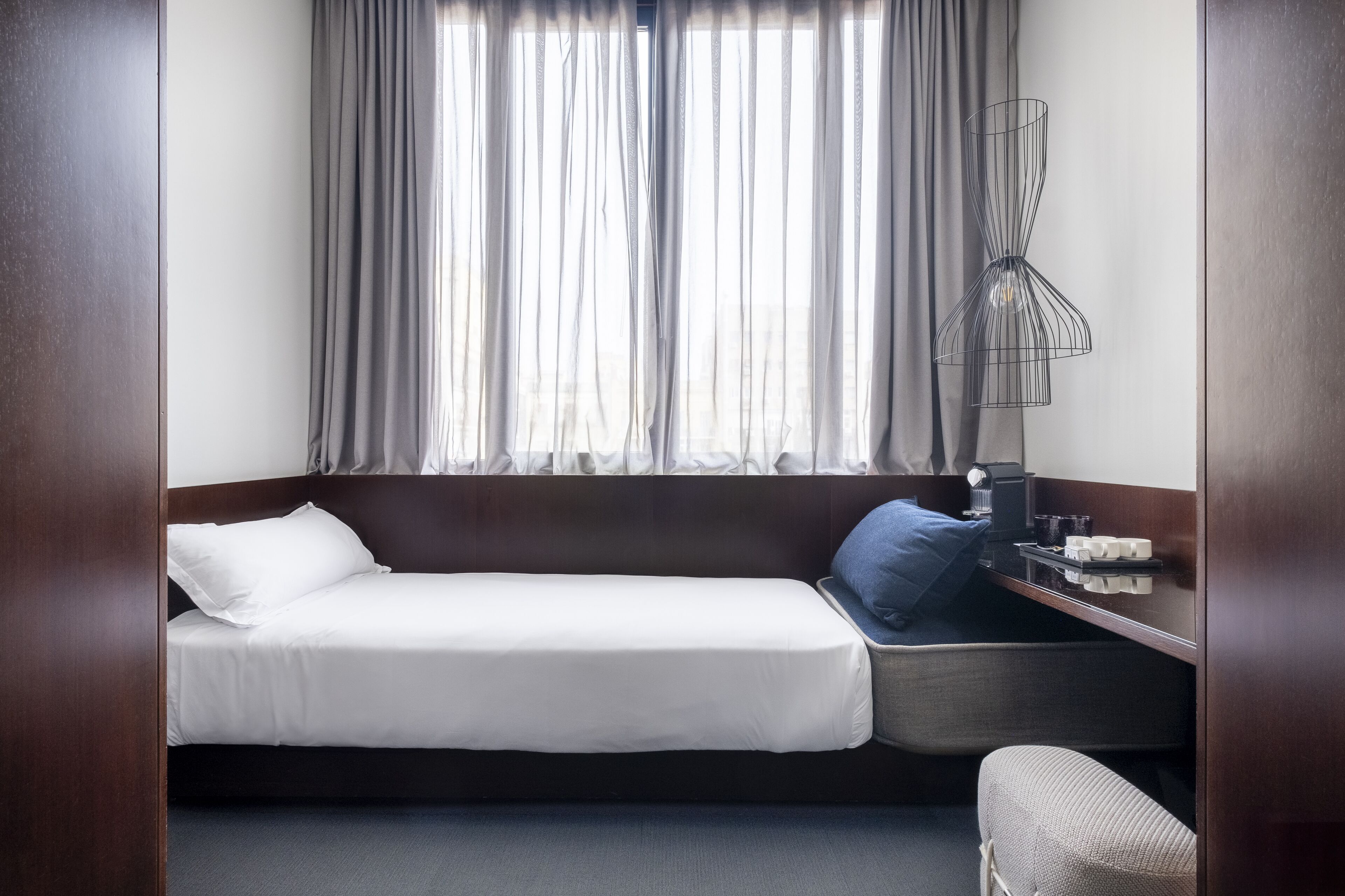 A hotel room with a single bed, a bedside table with a lamp, a chair, and a window with curtains.
