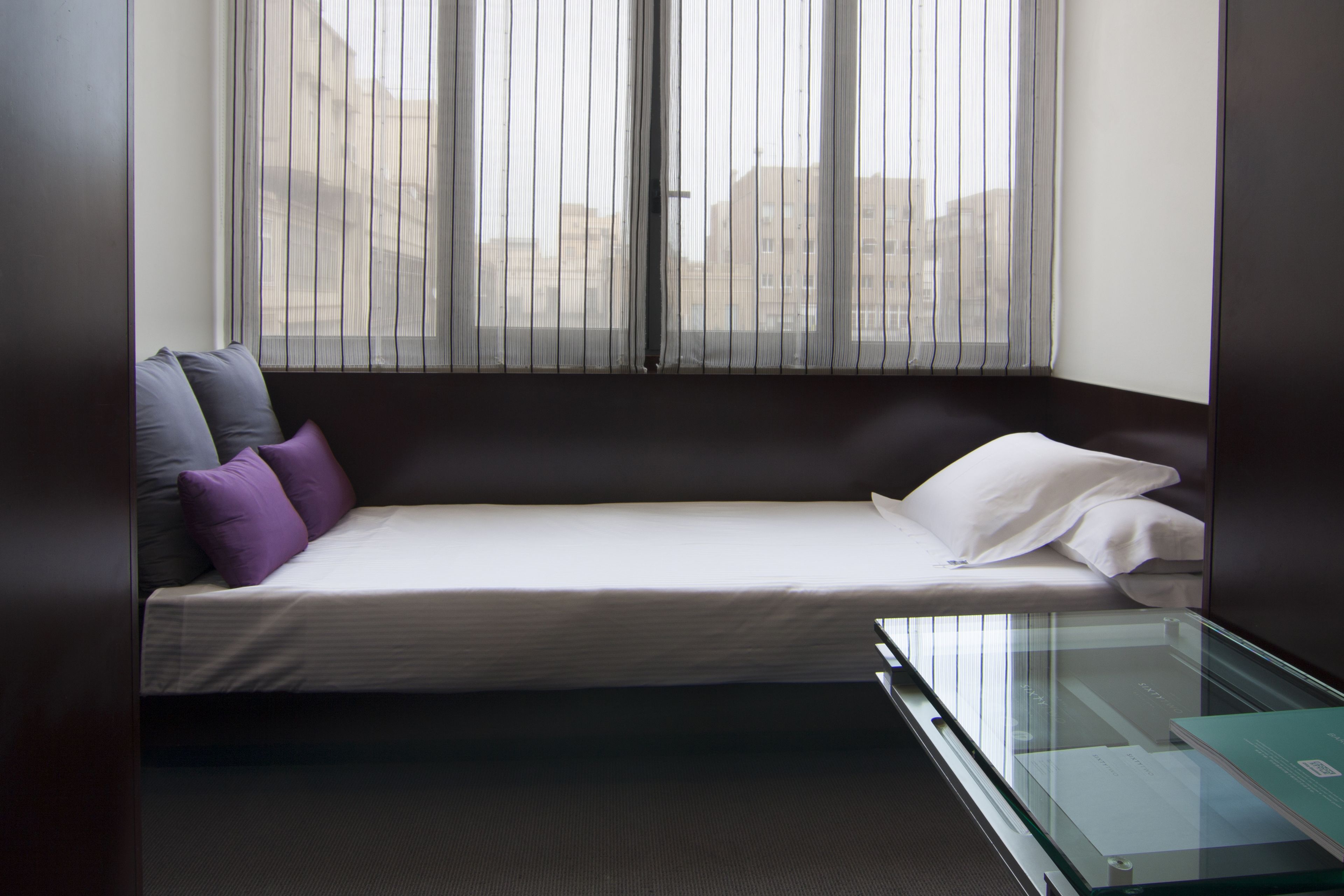Una habitación pequeña y moderna con una cama individual, una mesita de vidrio y vistas a través de cortinas traslúcidas.