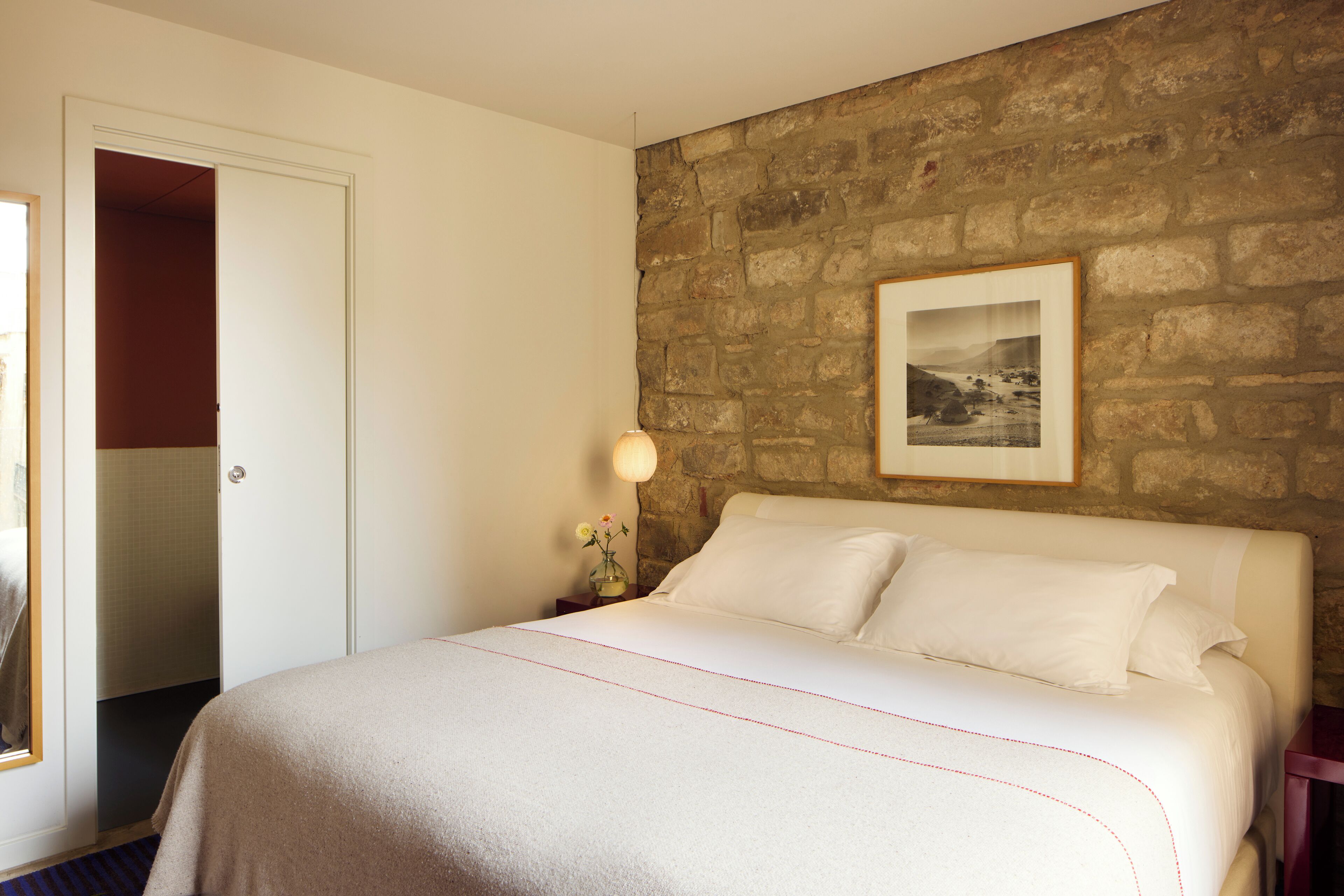 A bedroom with a stone wall, a framed picture, a bed with white linens, and a nightstand with a lamp and flowers.