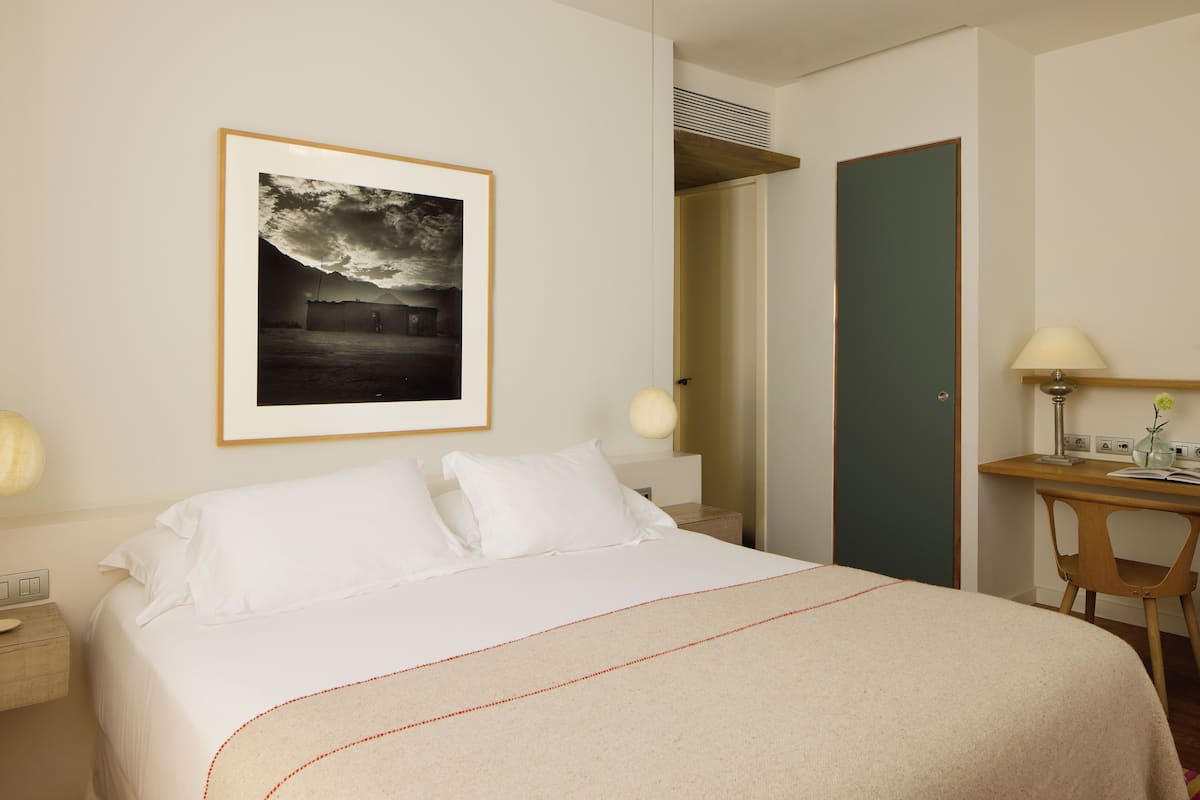 A hotel room with a large bed, a framed picture on the wall, a desk with a lamp, and a door leading to another room.