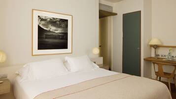 A hotel room with a large bed, a framed picture on the wall, a desk with a lamp, and a door leading to another room.