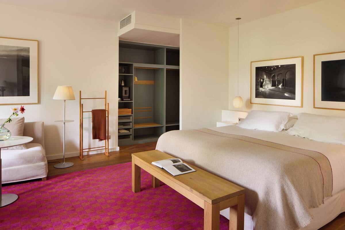 A hotel room with a bed, bedside tables, a bench, a desk, a chair, a wardrobe, and a large window.
