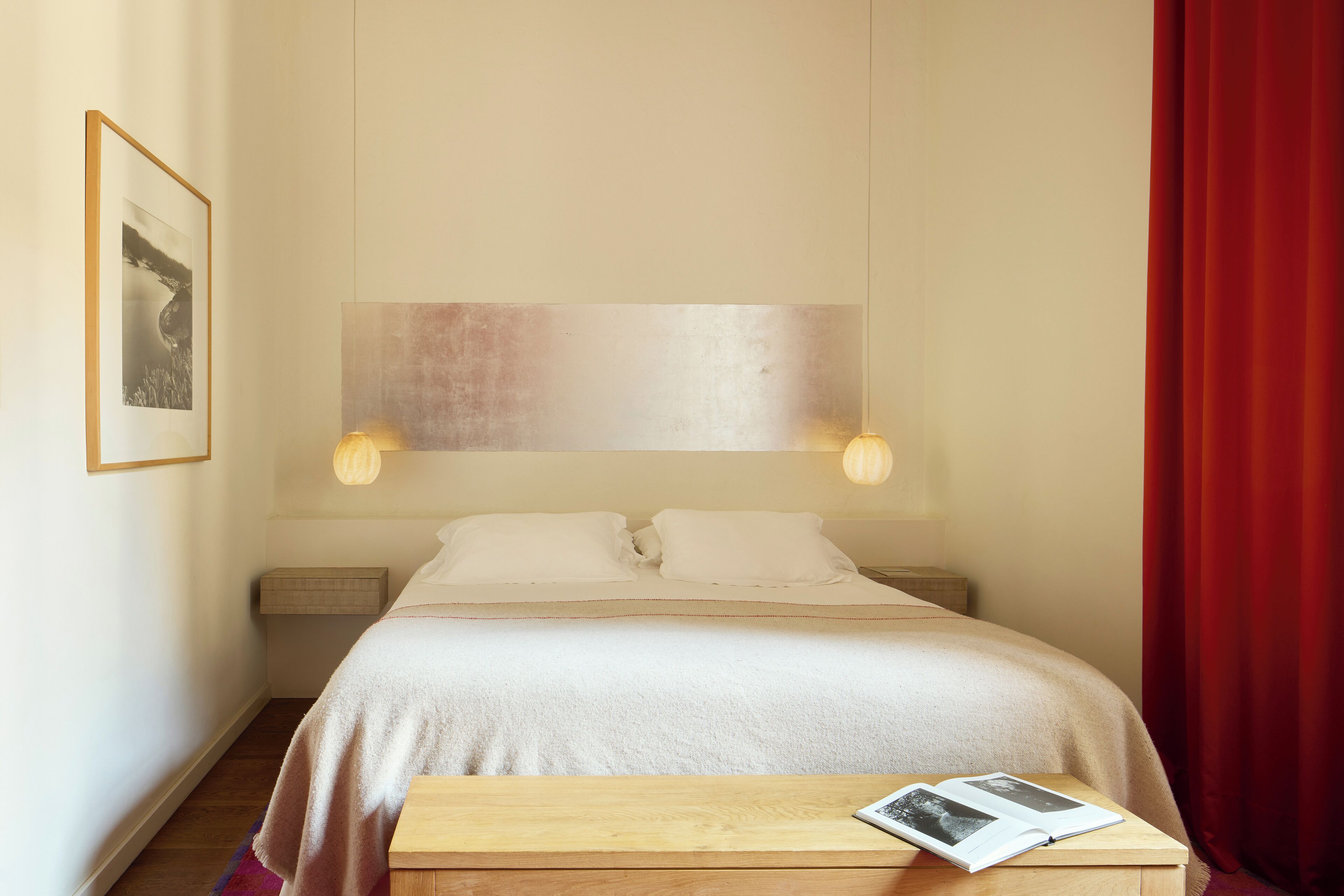 Quarto bem organizado com cama, mesas de cabeceira e uma pequena mesa de madeira com um livro.