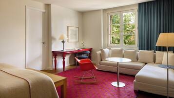 Habitación de hotel con cama, un sillón mecedora rojo, un sofá color beige, una mesita redonda pequeña, un escritorio con lámpara, y una ventana con vistas a árboles.