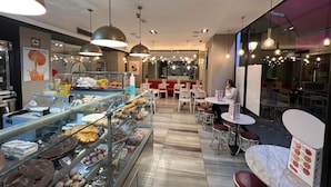 Una sección de panadería con una variedad de pasteles y postres exhibidos en una vitrina. Hay varias zonas de estar con mesas y sillas, y se ve a una persona sentada en una mesa.