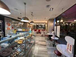 Ett avdelning i butiken med ett stort utbud av bakverk och desserter som visas upp i ett glasskåp. Det finns flera sittplatser med bord och stolar, och en person sitter vid ett bord.