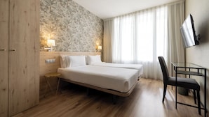 Habitación de hotel con cama doble, escritorio con silla, televisor montado en la pared y ventana con cortinas traslúcidas.