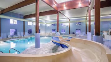 An indoor swimming pool with a slide, lounge chairs, and a view of the sky through windows.