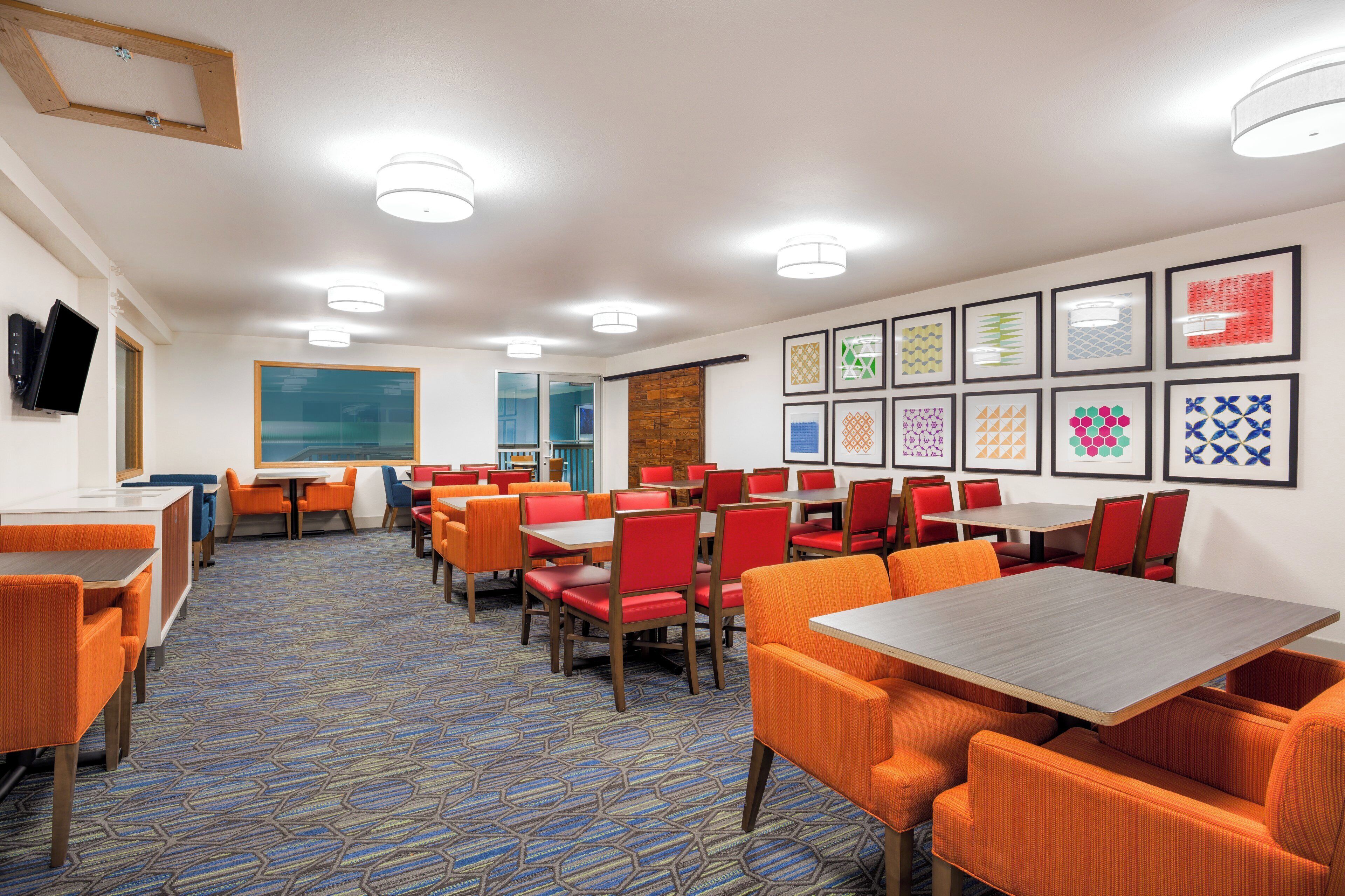 A hotel dining area with multiple tables and chairs, a patterned carpet, and framed artwork on the walls.