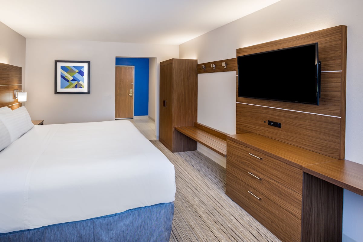 A hotel room with a large bed, a flat-screen TV mounted on a wooden cabinet, a framed artwork on the wall, and a blue door in the background.