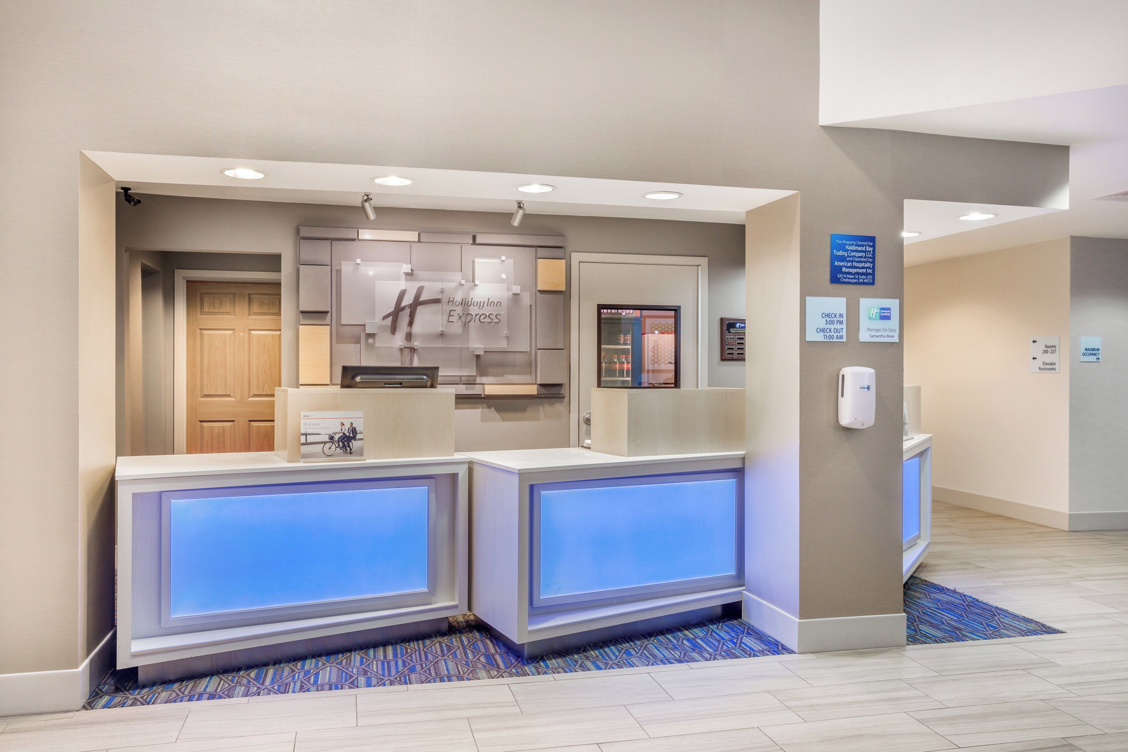 A hotel reception area with a modern design, featuring a reception desk, a digital display, and a sign indicating the property is managed by a hotel company.
