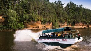 A boat with "Amazing Land and Water Dells Tours" written on the side, moving through a river surrounded by cliffs and trees. - Holiday Inn Express Wisconsin Dells by IHG (Baraboo)