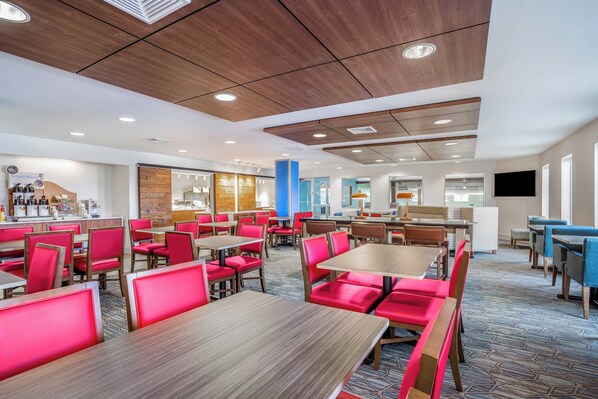 A modern dining area with wooden tables and red chairs, a kitchen area, and a seating area with blue chairs.