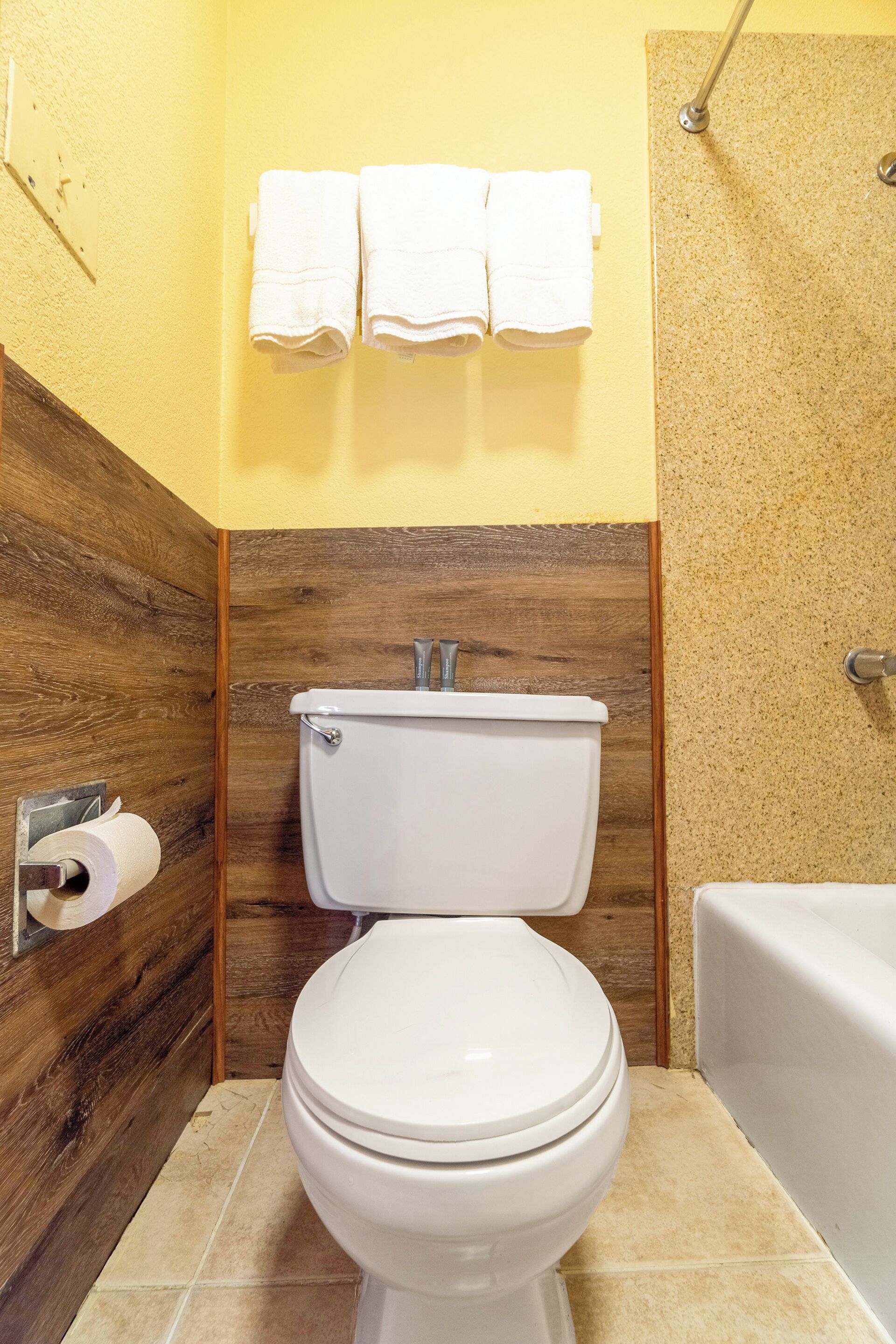 A bathroom with a toilet, a towel rack with three towels, a wooden wall, and a bathtub.