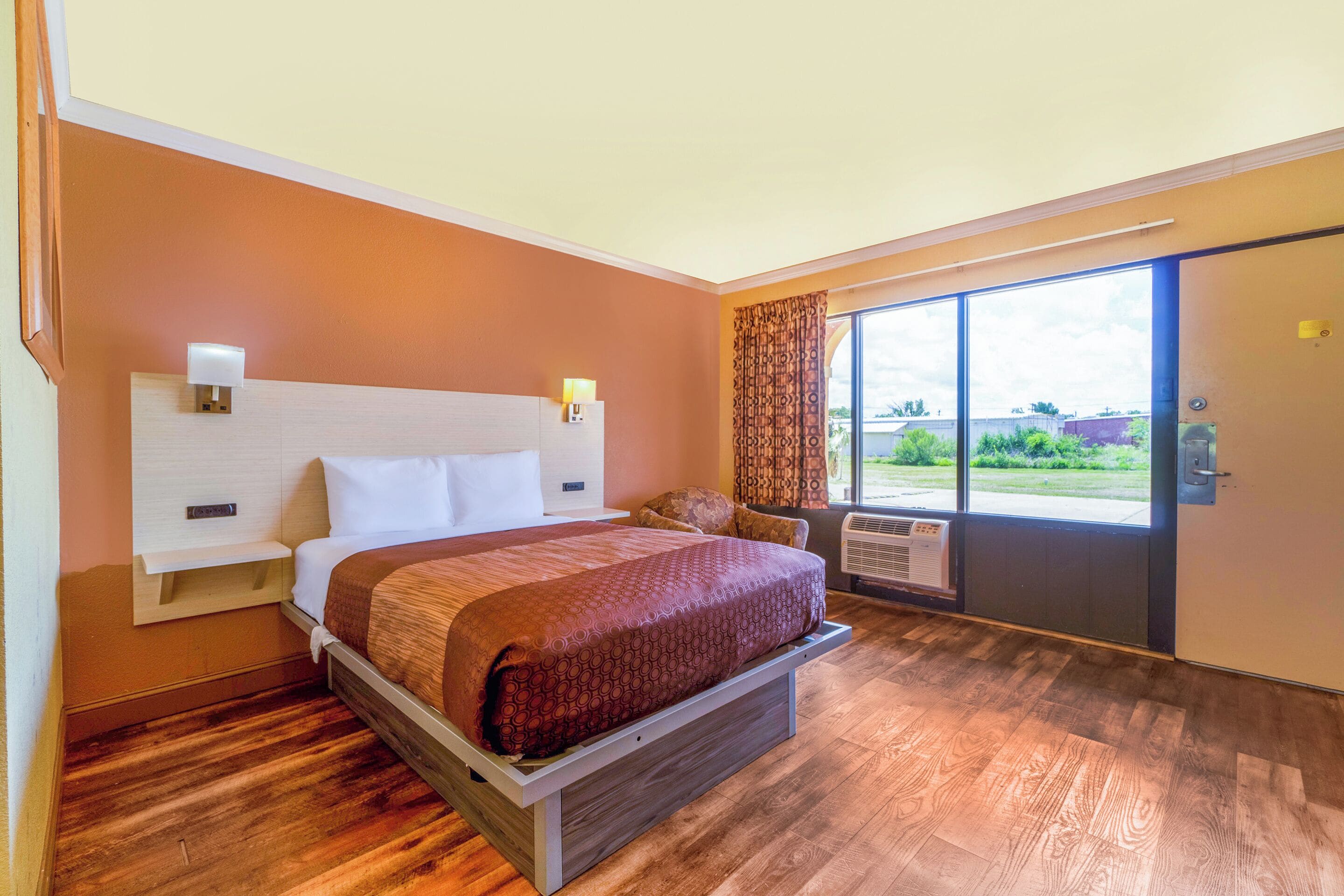 A hotel room with a bed, a nightstand, a window with curtains, and an air conditioning unit.