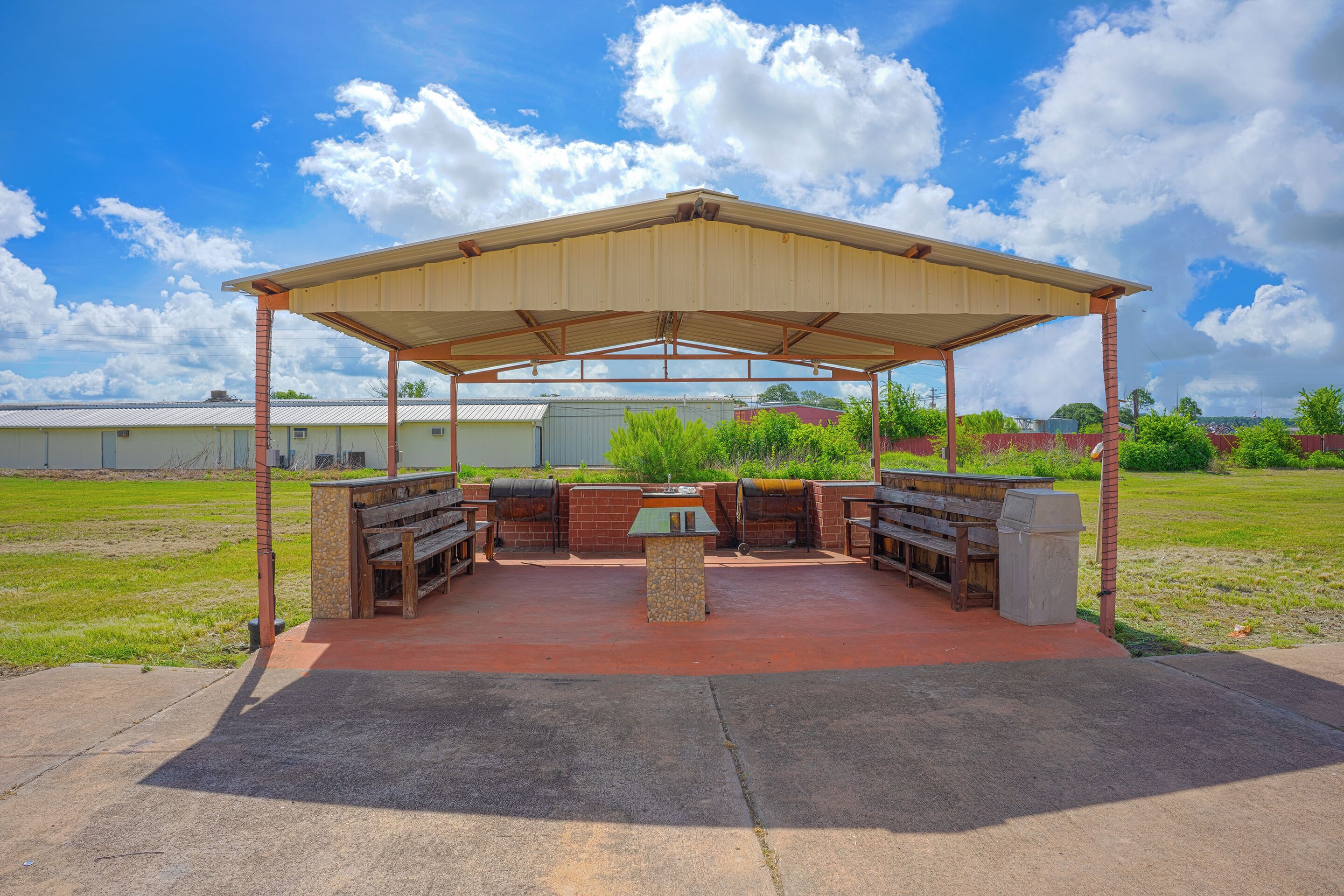 Un patio al aire libre con un área de estar cubierta y una mesa central, rodeado por un campo de césped y edificios industriales al fondo.