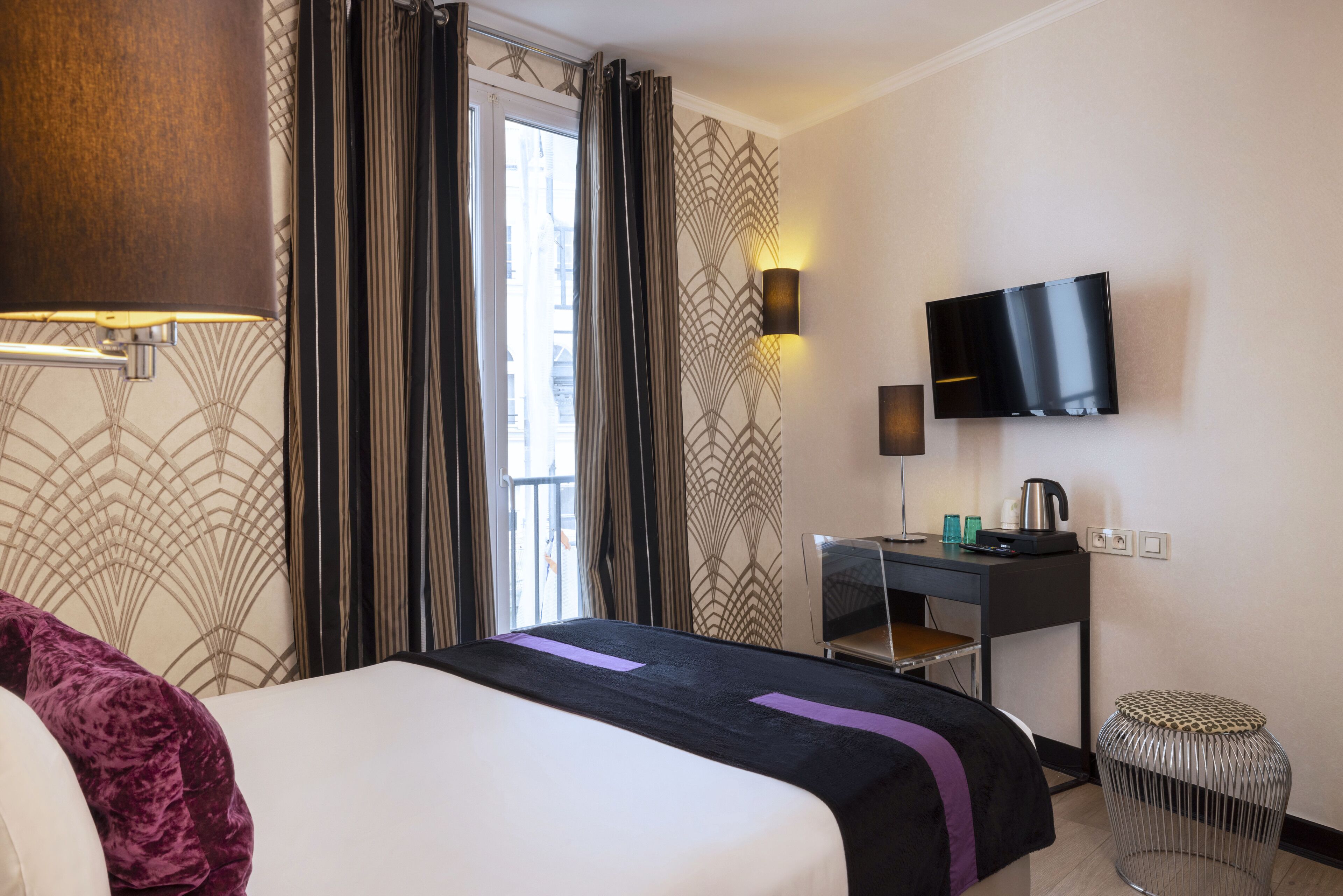 A hotel room with a bed, a desk with a flat-screen TV, a kettle, and a small stool.
