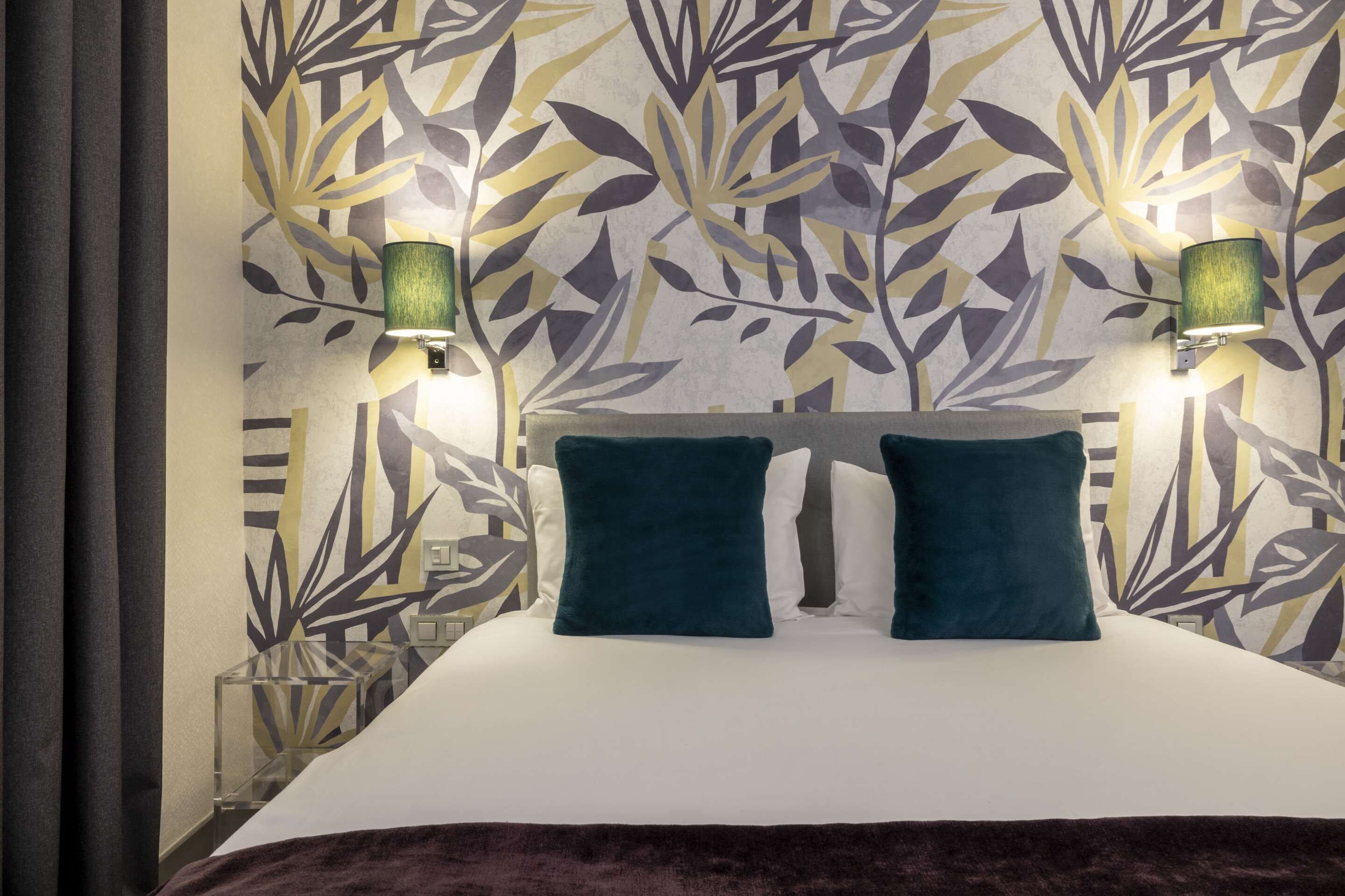 Une chambre d’hôtel avec un lit, deux oreillers, une table de chevet et un mur orné d’un motif de feuilles.