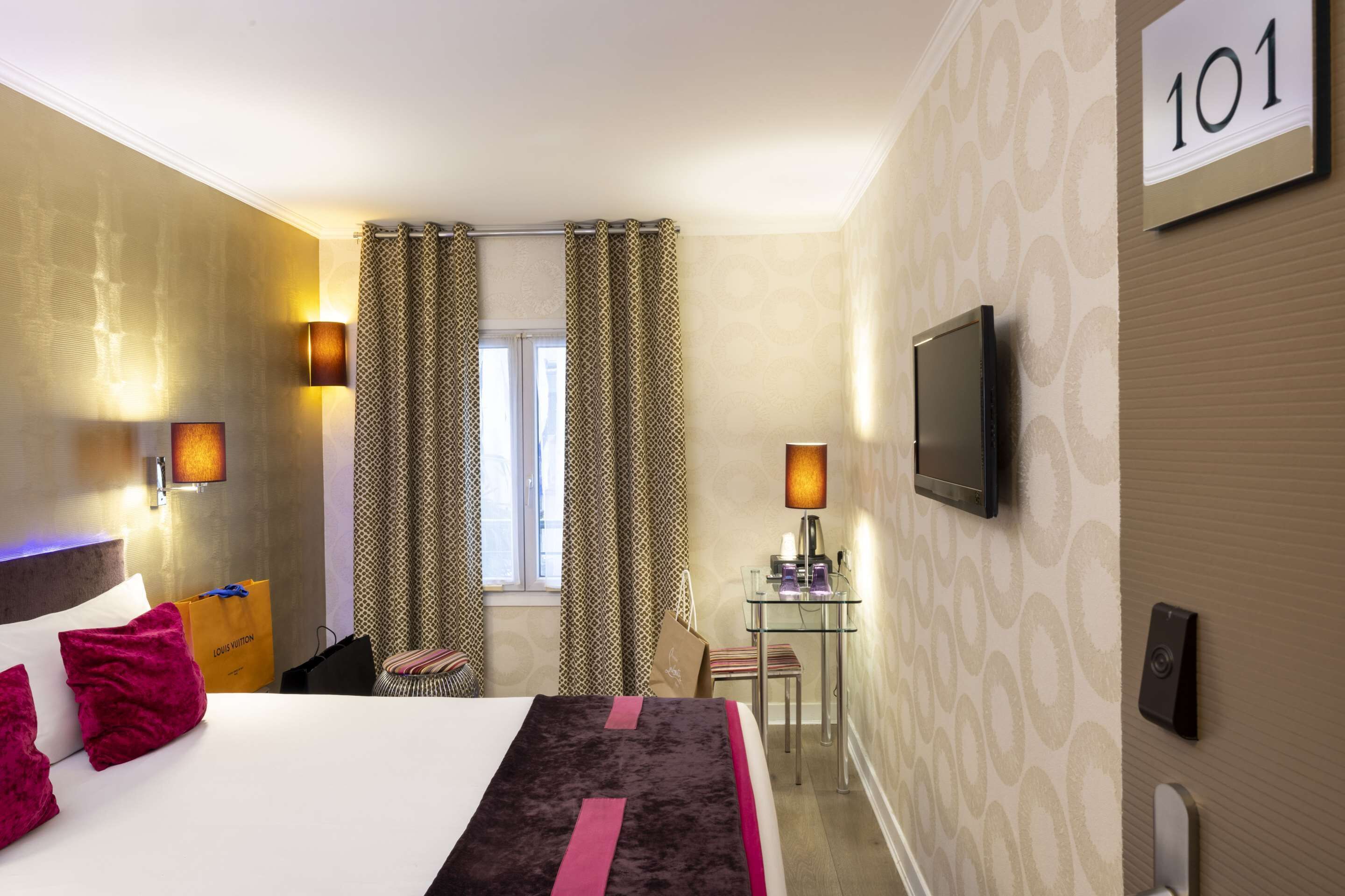 Habitación de hotel con una cama grande, dos lámparas de noche, un escritorio con silla, un televisor montado en la pared y una ventana con cortinas.