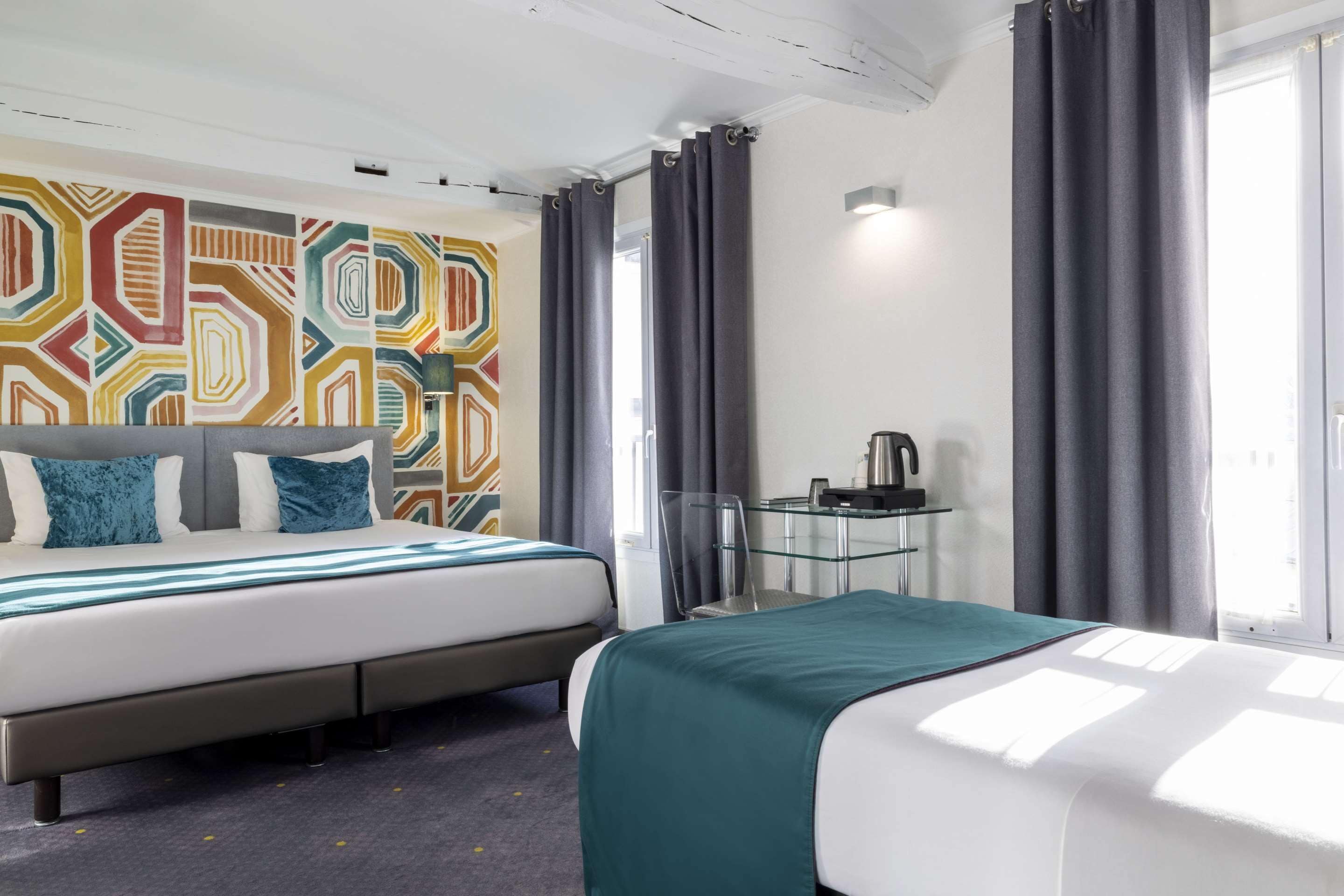 Ein Hotelzimmer mit zwei Betten, einem Glastisch, einer Kaffeemaschine und einem farbenfrohen, geometrischen Wanddesign.