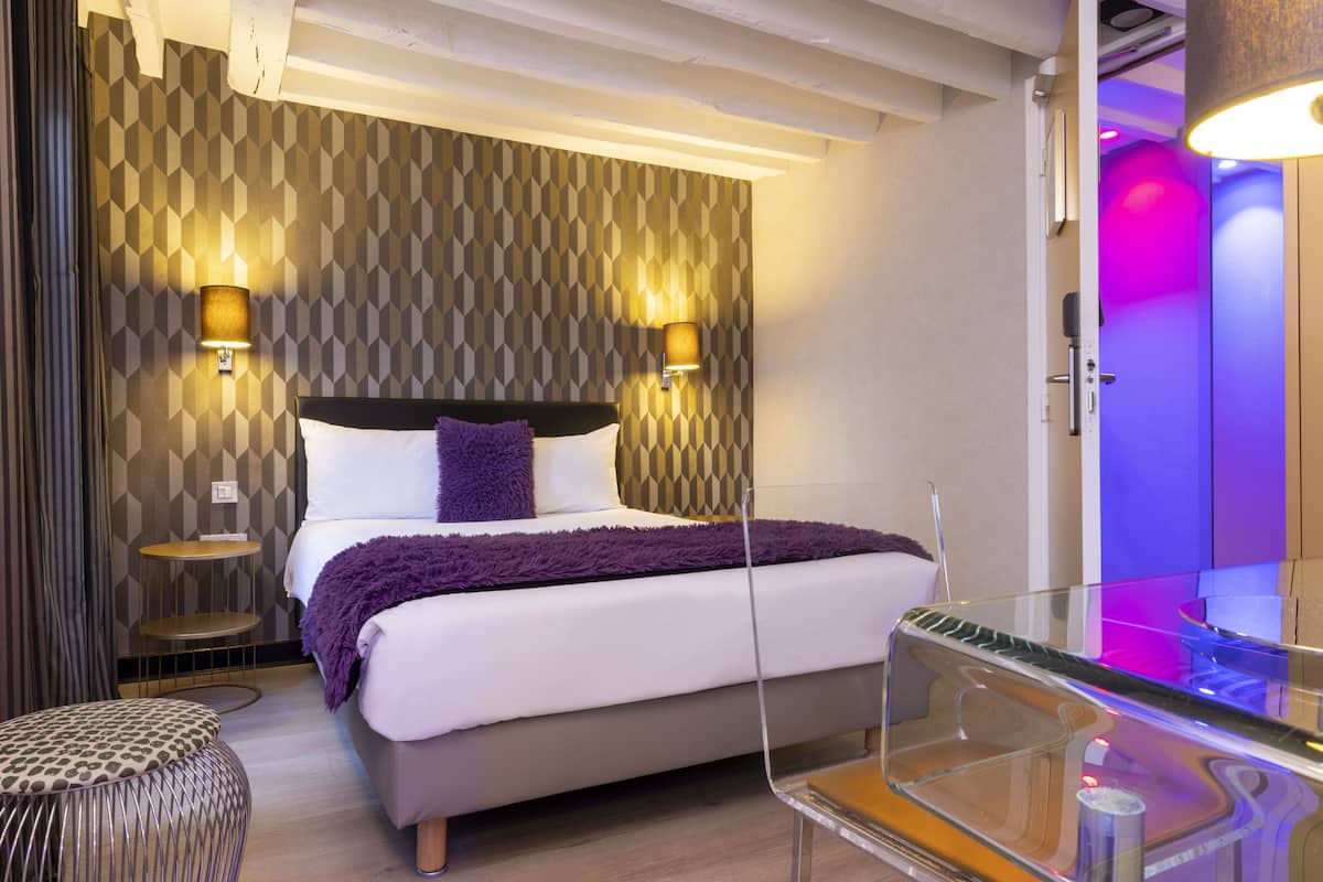 Een moderne hotelkamer met een bed, twee wandlampen, een rond poefje en een glazen tafeltje.