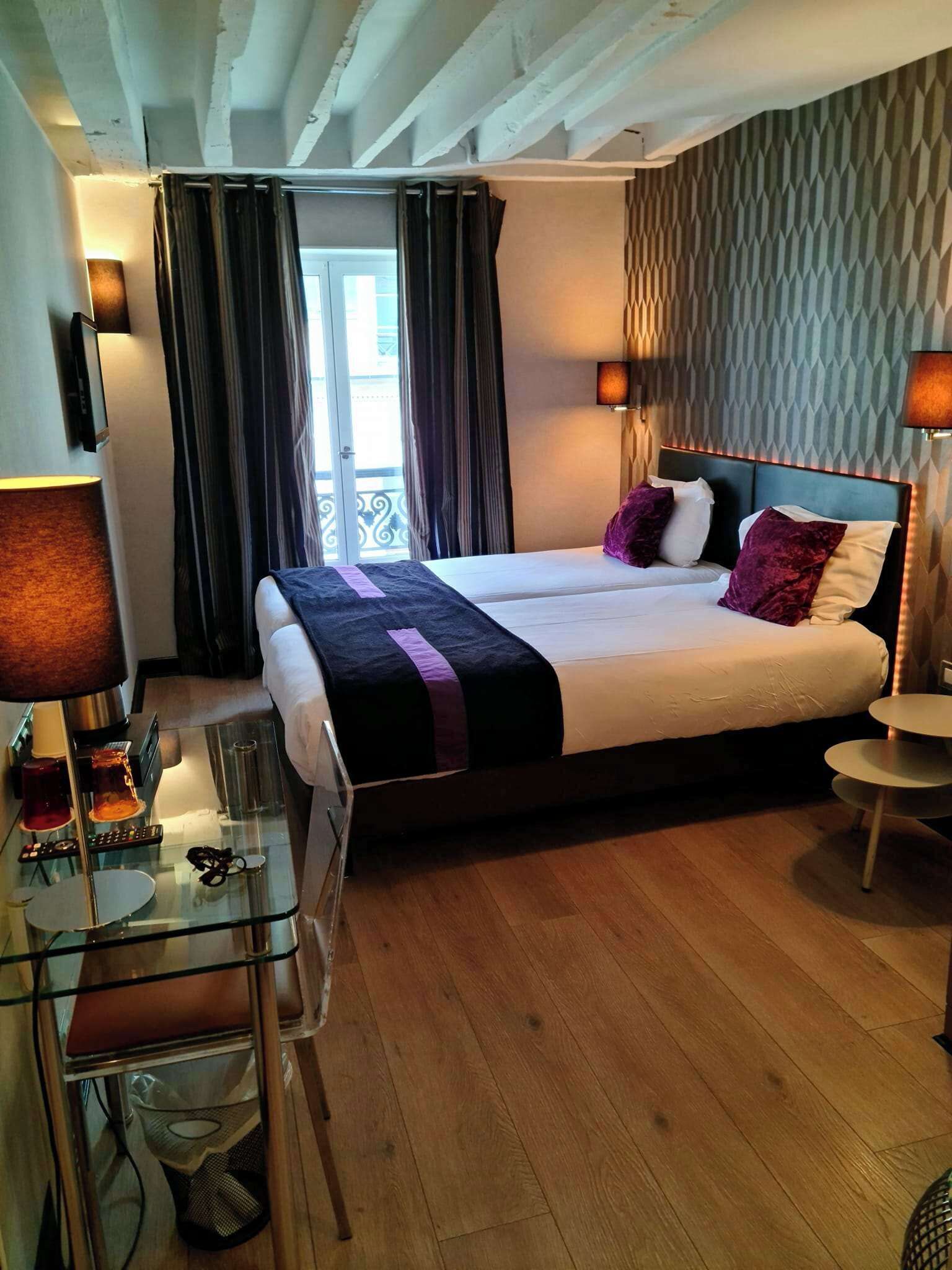 A hotel room with a large bed, a glass desk, a small round table, and a balcony with curtains.