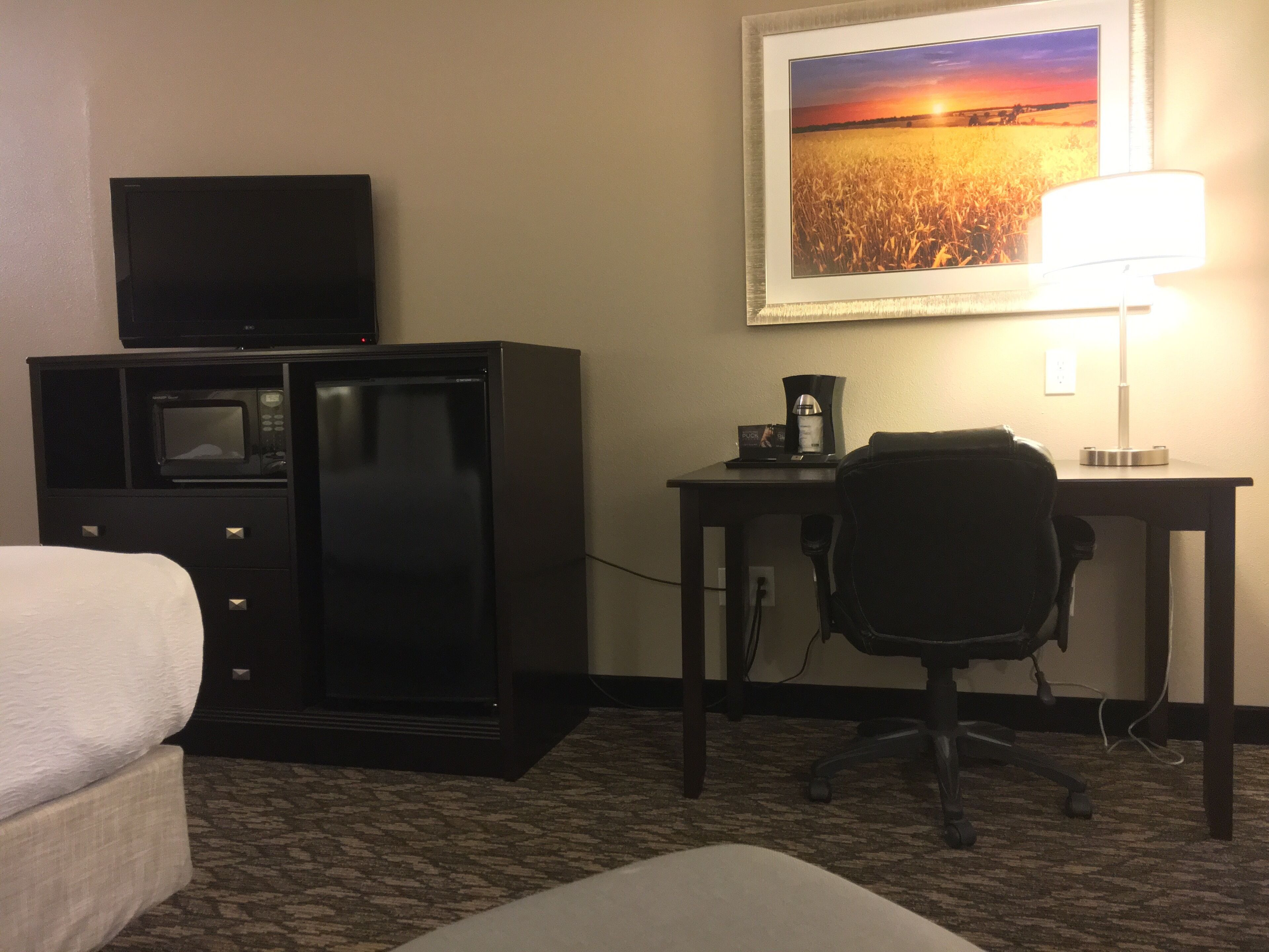 A hotel room with a bed, a desk with a lamp, a microwave, a coffee maker, and a framed picture on the wall.
