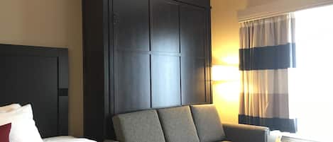 Une chambre d’hôtel avec un grand lit, un canapé, une armoire en bois foncé, un téléviseur fixé au mur et une fenêtre avec des rideaux.