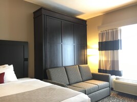 A hotel room with a large bed, a sofa, a dark wooden wardrobe, a wall-mounted TV, and a window with curtains.