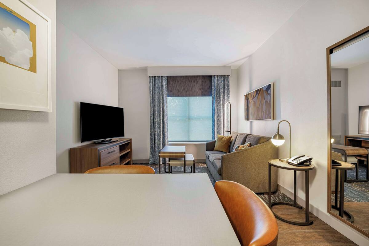 A hotel room with a flat-screen TV, a dining table, a sofa, a small side table with a lamp, and a large mirror.