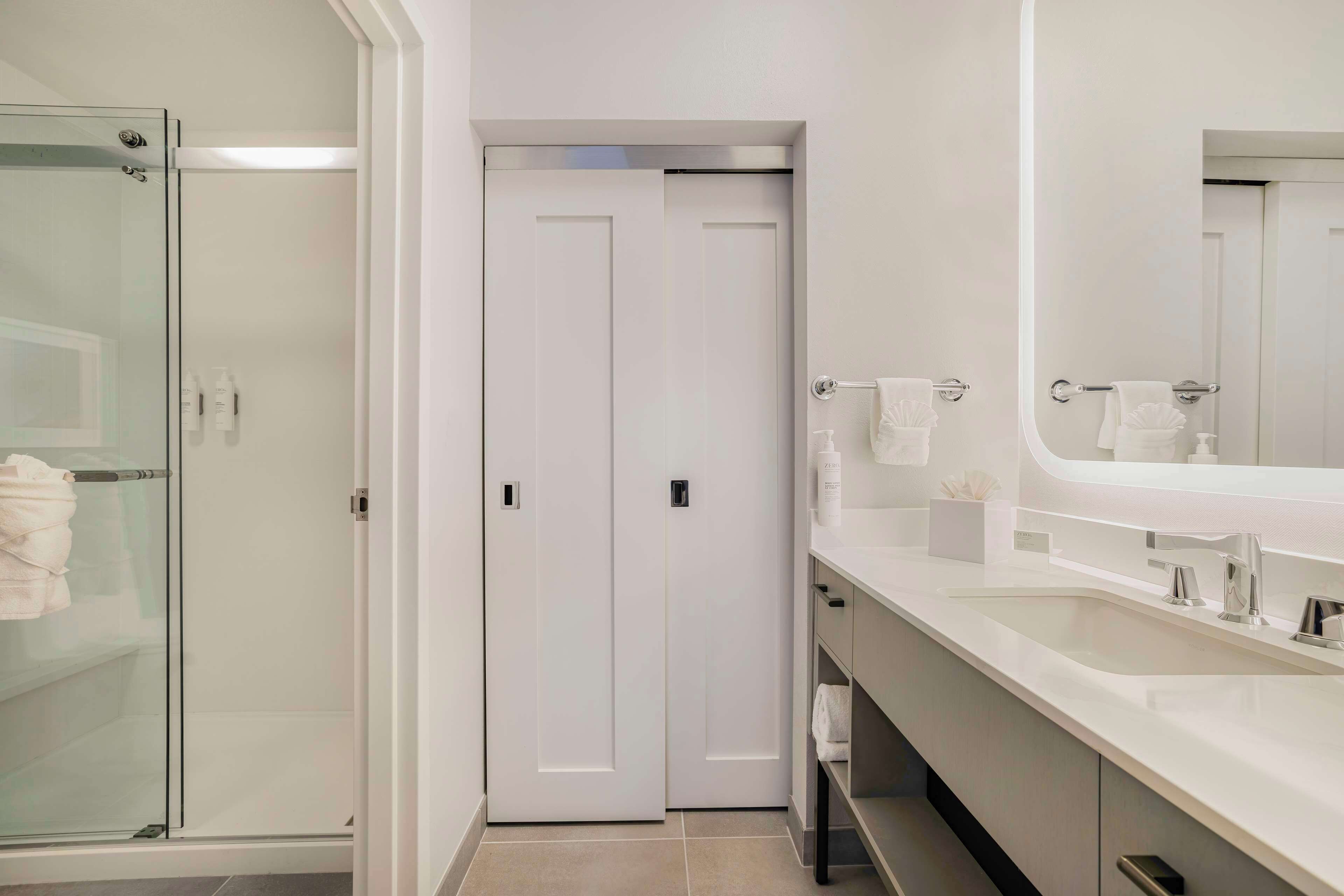 A modern bathroom with a glass shower enclosure, a double door, a large mirror, and a white countertop with a sink and vanity.