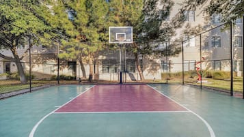 An outdoor basketball court with a chain-link fence, trees, and a building in the background.