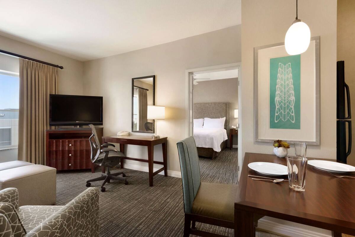 A hotel room with a TV, desk, chair, dining table with plates, a mirror, and a bed.