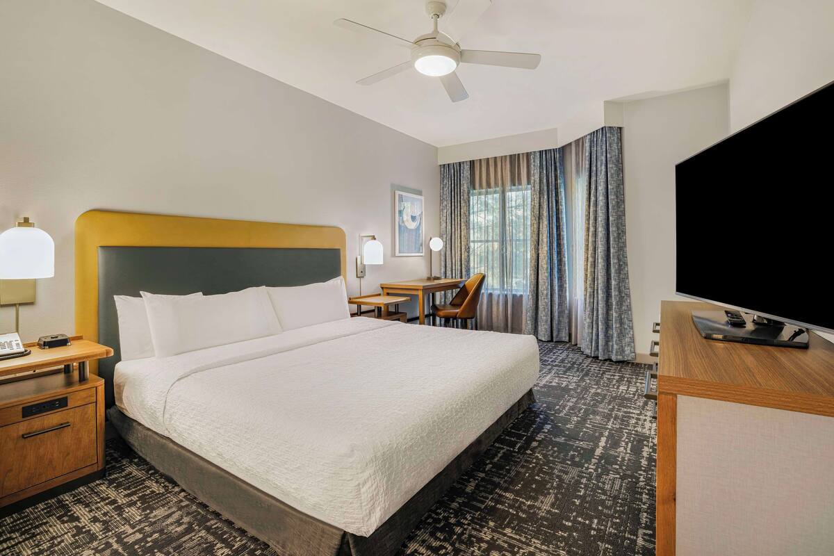 A hotel room with a large bed, a desk with a lamp, a television, and a ceiling fan.
