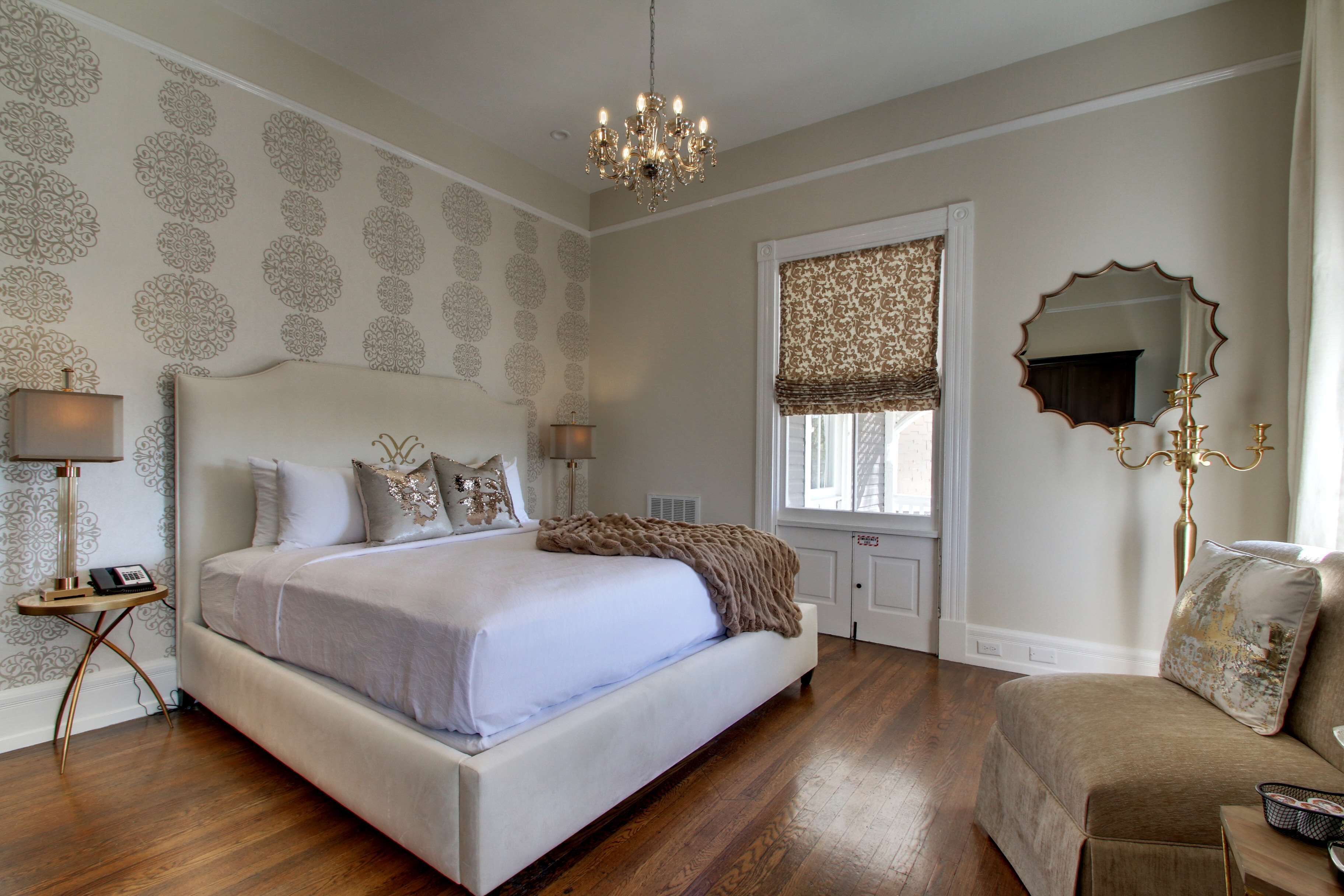 A bedroom with a large bed, a chandelier, a window with patterned curtains, and a mirror on the wall.