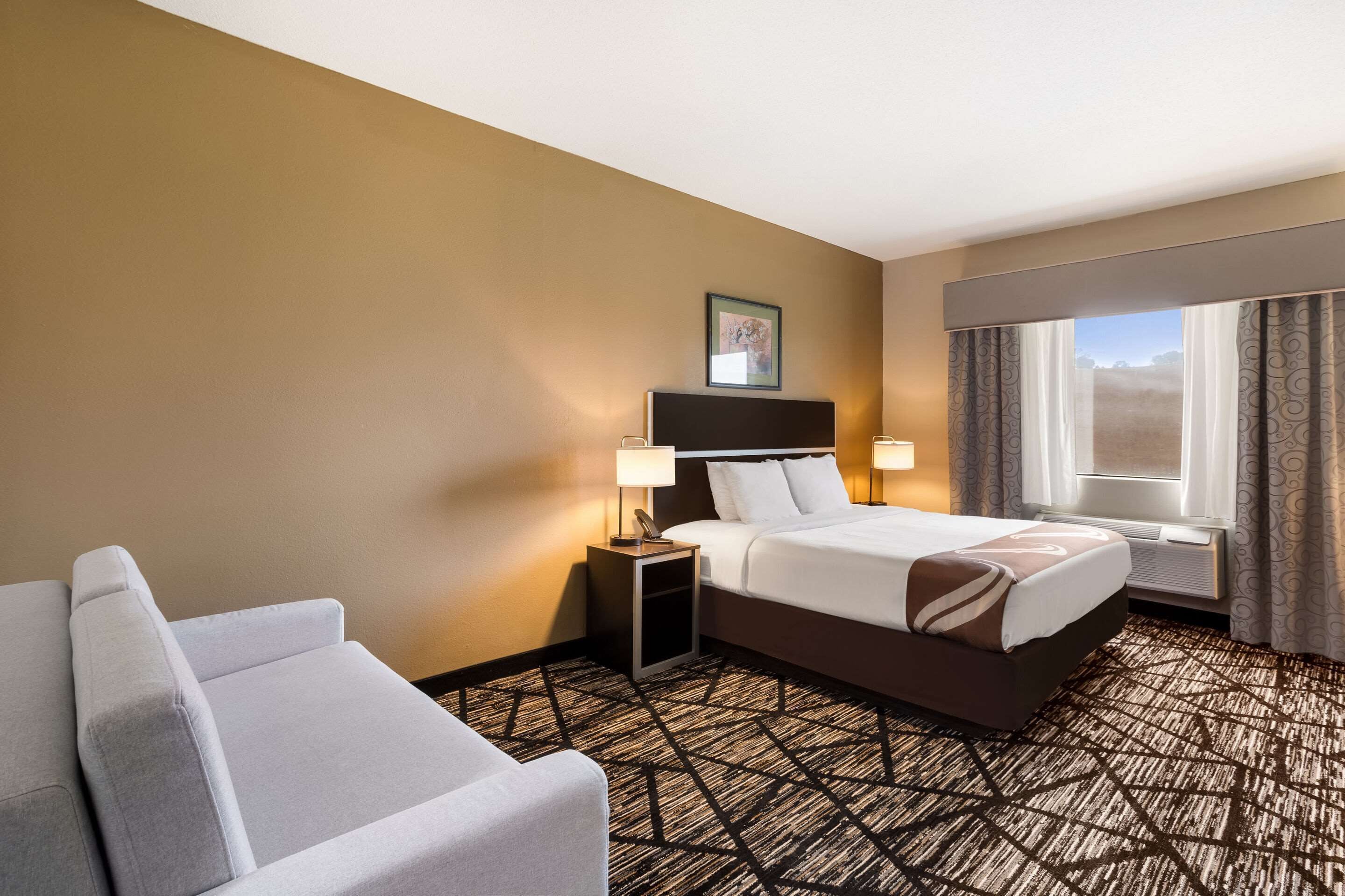 Une chambre d’hôtel comprenant un lit, une table de chevet avec des lampes, une chaise, une fenêtre avec des rideaux et un tapis à motifs géométriques.