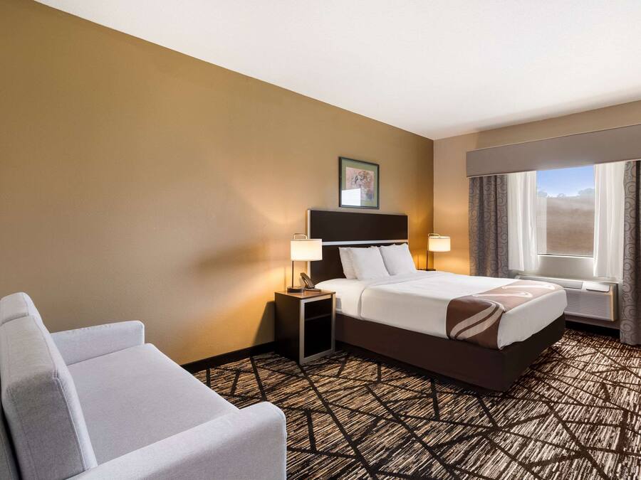 Habitación de hotel con cama, mesita de noche con lámparas, una silla, ventana con cortinas y alfombra con diseño geométrico.