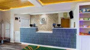 A hotel reception area with a herringbone-patterned wall, a reception desk with two monitors, a digital screen, and a small shelf with snacks. - Quality Inn & Suites Pearl - Jackson (Pearl)