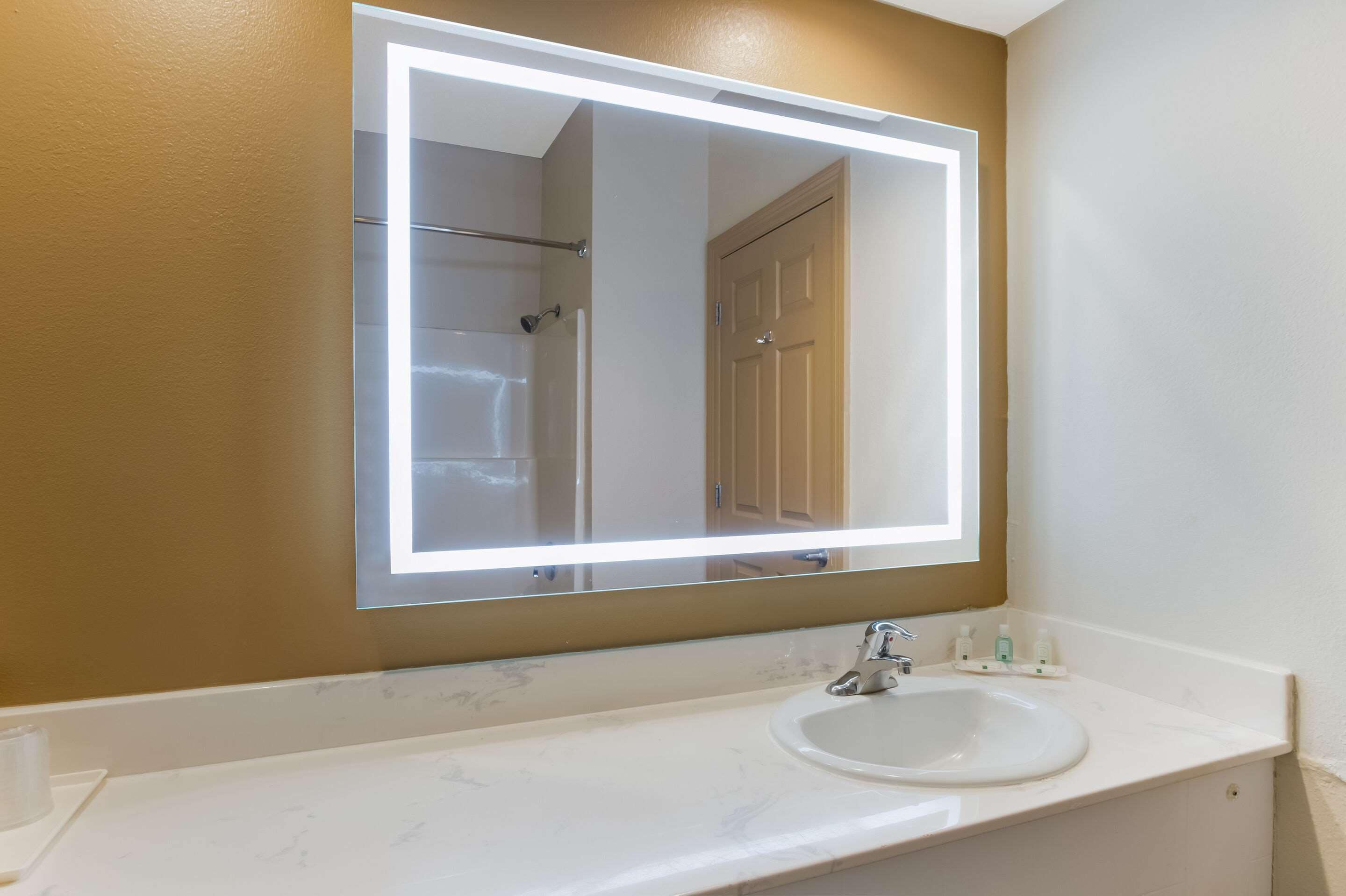 Une salle de bain avec un lavabo blanc, un miroir doté d’un cadre éclairé et une porte.
