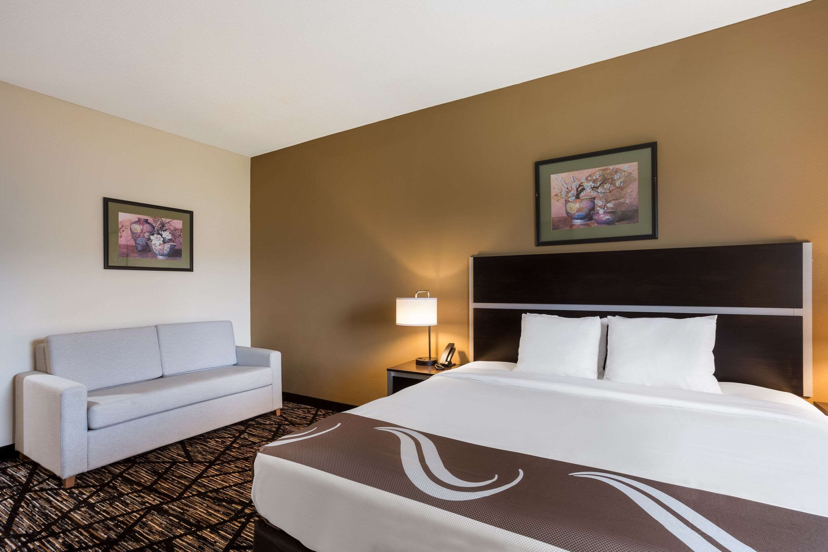 Ein Hotelzimmer mit einem großen Bett, einem Sofa, einem Nachttisch mit Lampe und zwei gerahmten Bildern an der Wand.