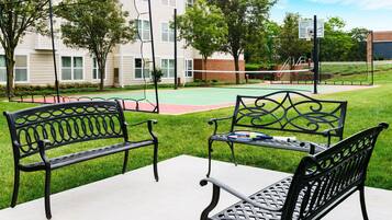 Un espacio de estar al aire libre con dos bancos de metal negro y una mesa, una cancha de baloncesto y un edificio con ventanas al fondo.