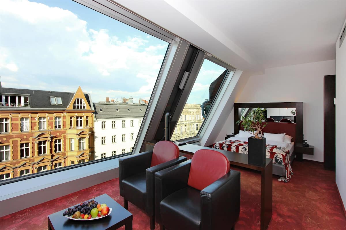 A modern hotel room with a large window, a bed, a desk, and a dining area with fruit.